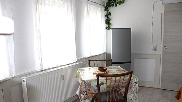 A dining area with a table covered by a floral tablecloth, wooden chairs, a bowl on the table, and a radiator below the window.