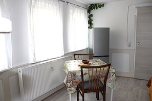 Ein Essbereich mit einem Tisch, der mit einer floralen Tischdecke bedeckt ist, Holzstühle, eine Schüssel auf dem Tisch und einem Heizkörper unter dem Fenster.