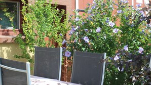 Un área de estar al aire libre con una mesa y sillas, rodeada de un jardín con flores púrpuras.