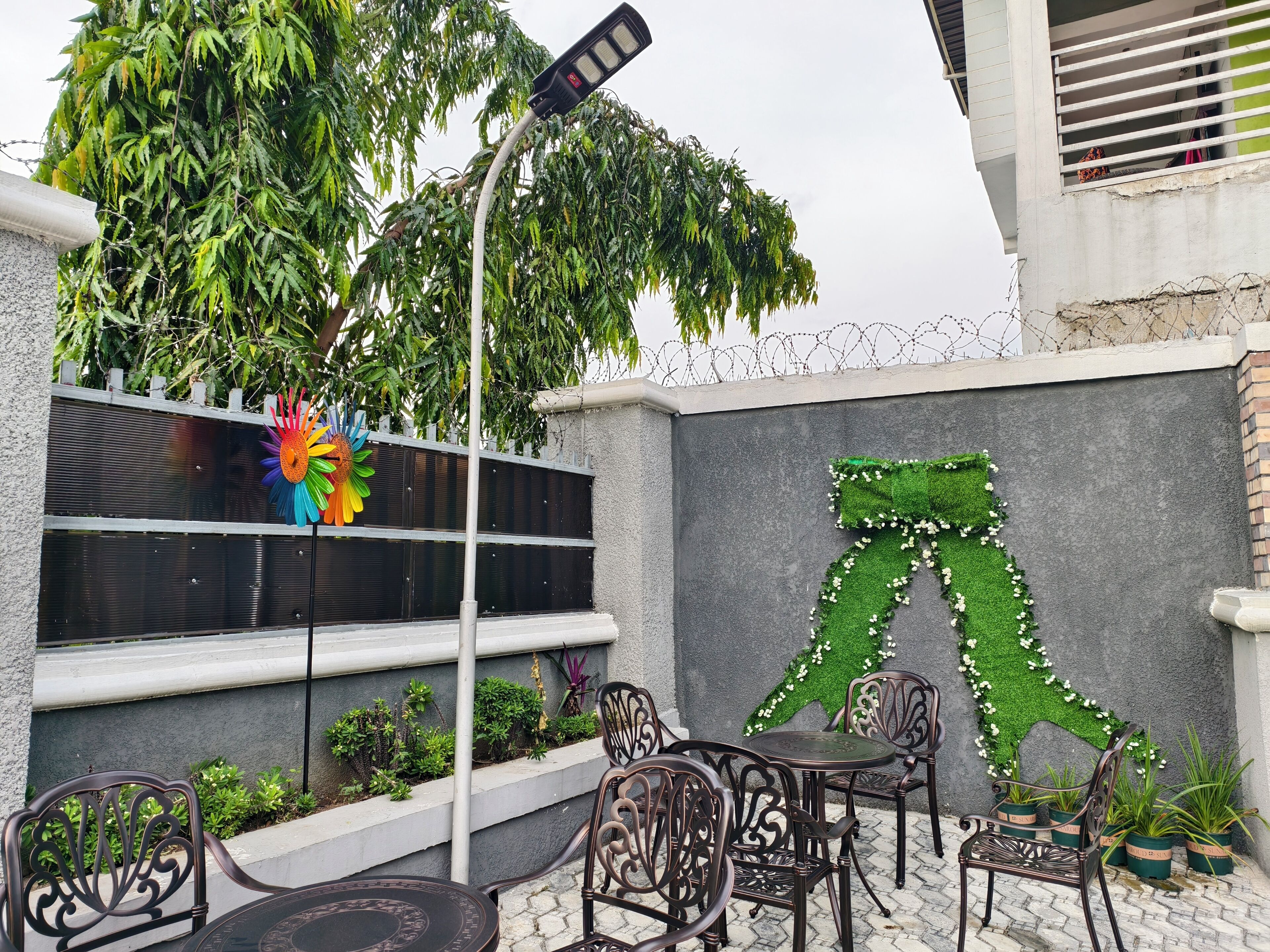 An outdoor seating area with metal chairs and a table, surrounded by potted plants and a decorative wall with a green design.