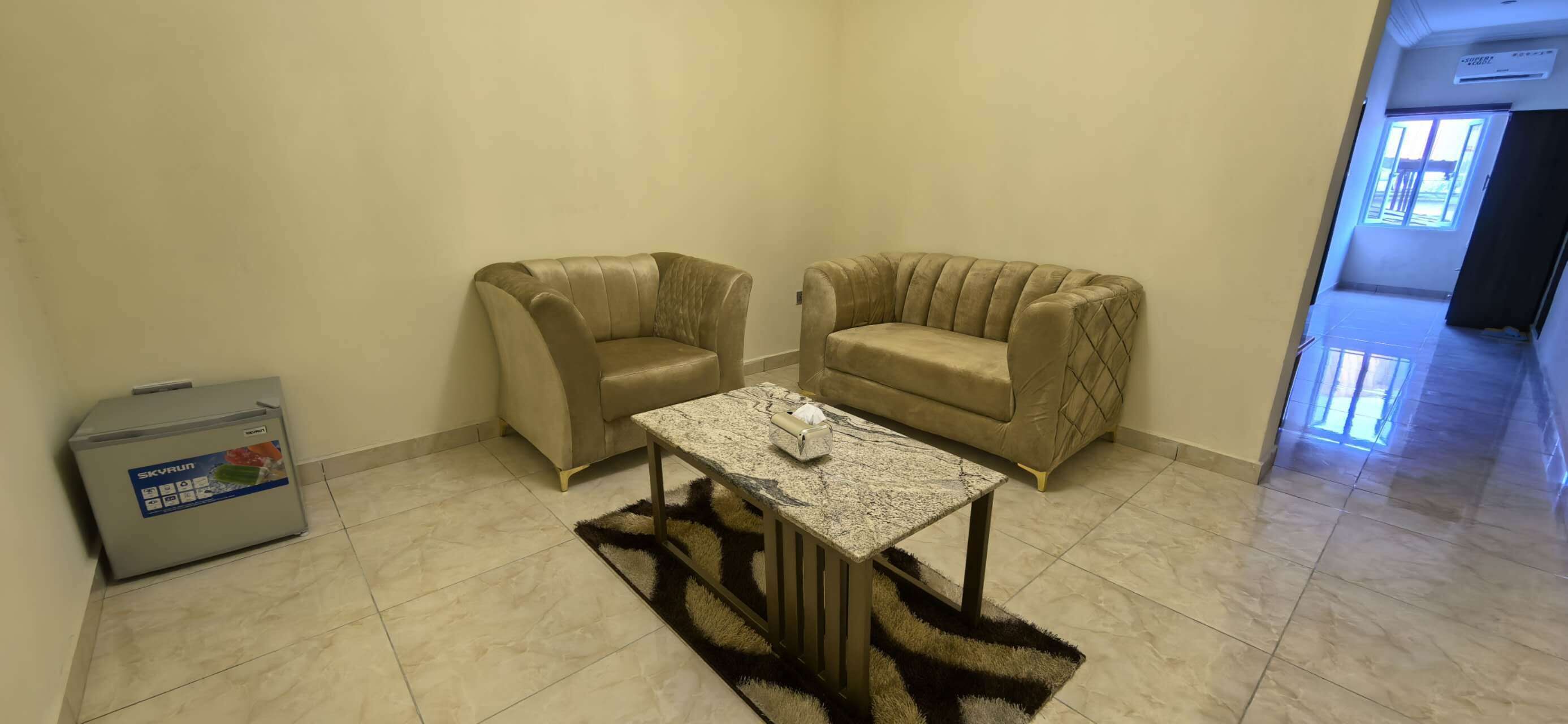A room with two beige armchairs, a small table with a decorative item, and a refrigerator.