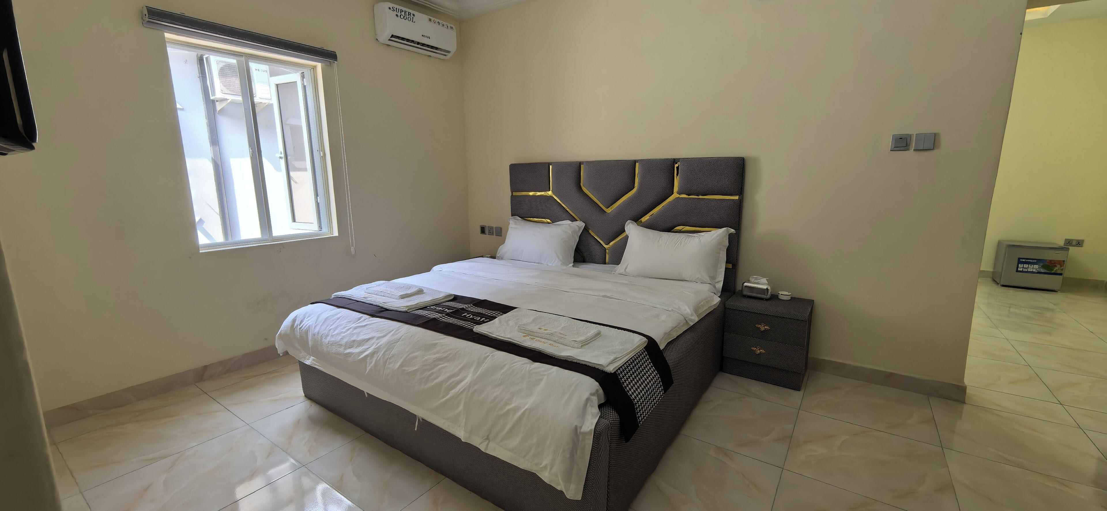 A neatly made bed with a headboard, a bedside table with a lamp, and a window with blinds.
