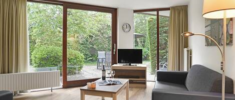 A modern living room with a television, a sofa, a coffee table, and a lamp. There is a large window with curtains, a clock on the wall, and a view of greenery outside.