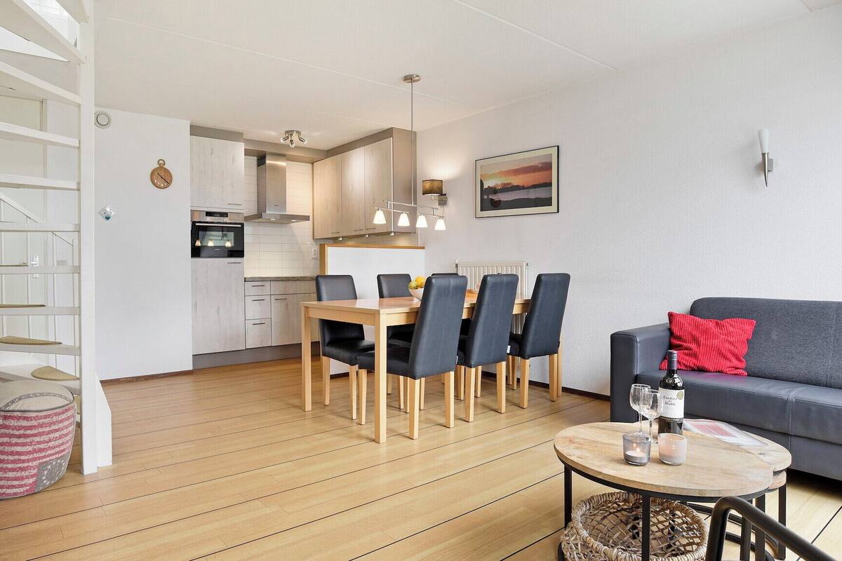 Un saló modern amb un sofà gris, una taula de dinar amb cadires, una zona de cuina i una escala amb un televisor a la paret.