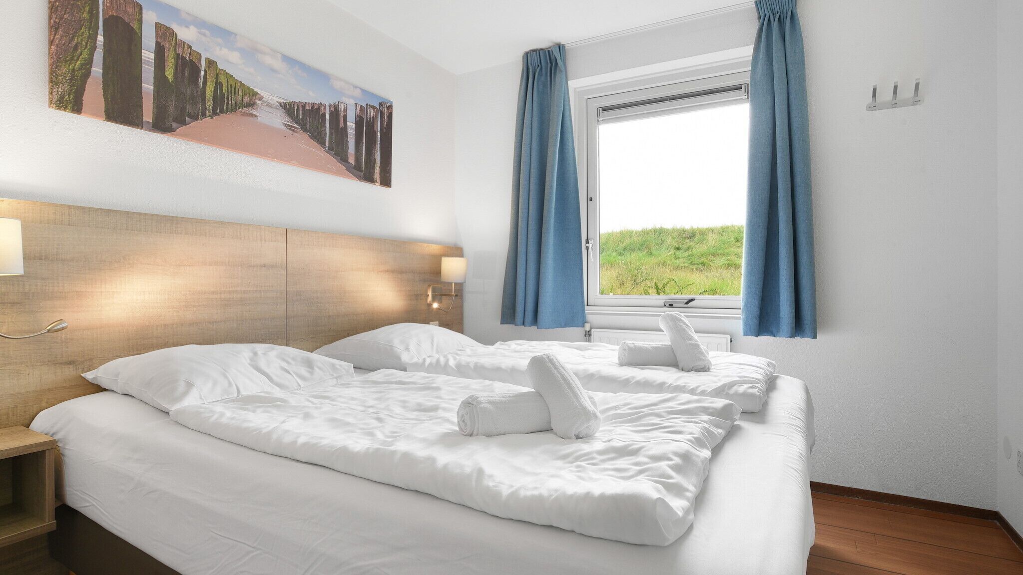 Une chambre d’hôtel avec un grand lit, une tête de lit dotée d’un luminaire, une fenêtre avec des rideaux et un tableau au-dessus du lit.