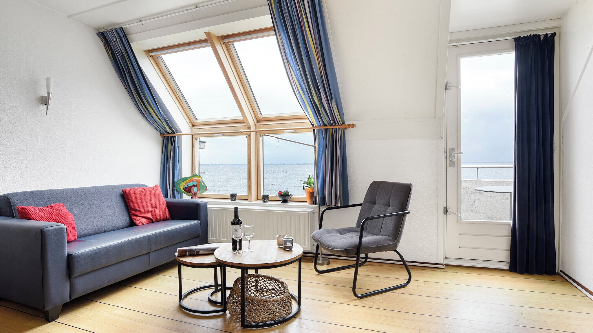 A room with a blue sofa, a grey armchair, a small round table, and a view of the sea through a large window.