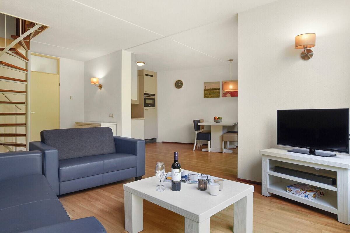 Un saló modern amb un sofà, una taula de centre amb una ampolla, gots i un comandament a distància. Hi ha una televisió sobre un moble baix i una escala al fons.
