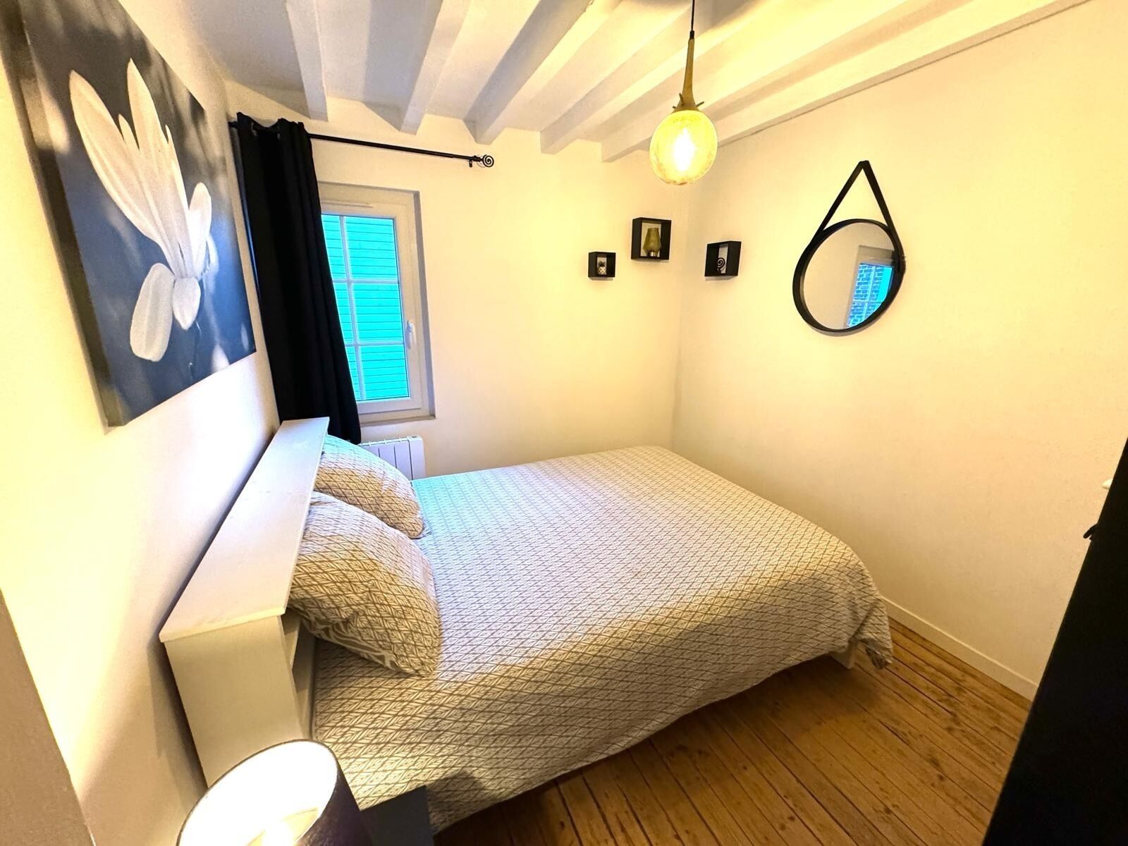 A small, neatly arranged bedroom with a bed, a window with blinds, a wall-mounted mirror, and a ceiling light fixture.
