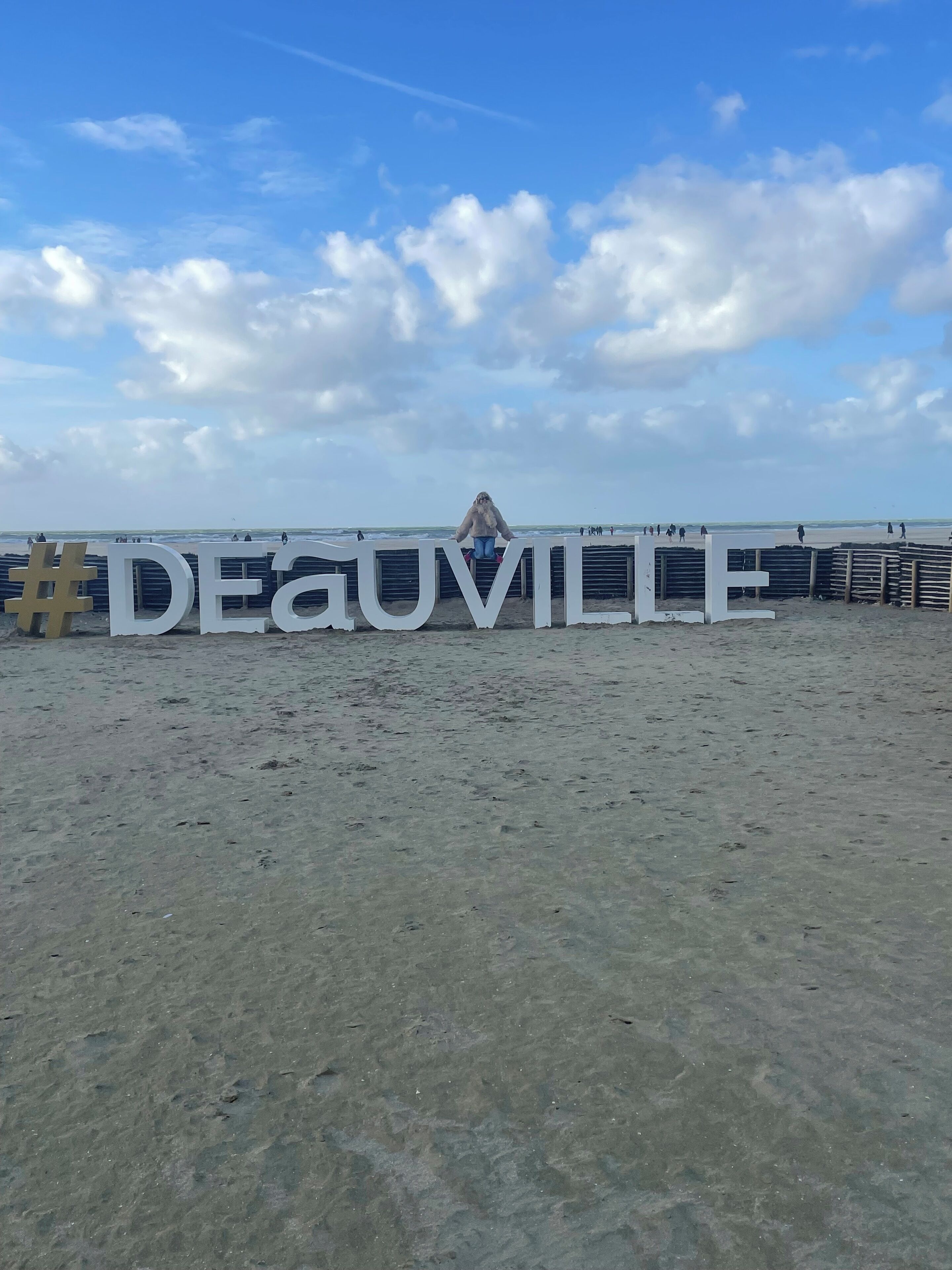 Ein Strand mit einem großen, weißen Schild, auf dem "DEAUVILLE" steht, und einem Holzzaun im Hintergrund.