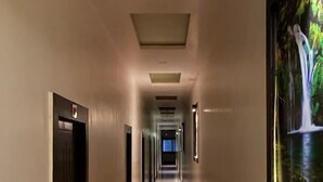 Ein langer, schmaler Korridor mit Türen an beiden Seiten, eingelassener Beleuchtung und einem großen, gerahmten Bild an der rechten Wand.
