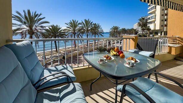 A balcony with a glass table, blue cushioned seating, and a view of the beach and palm trees.