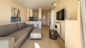 A modern living room with a grey sofa, a small ottoman, a flat-screen TV mounted on the wall, and a kitchen area in the background.
