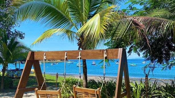 Un columpio de madera con dos columpios, una palmera y vista al mar con barcos.