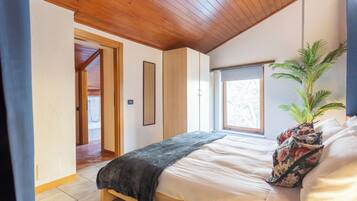 A bedroom with a wooden ceiling, a bed with white bedding, a dark blanket, a floral pillow, and a potted plant.