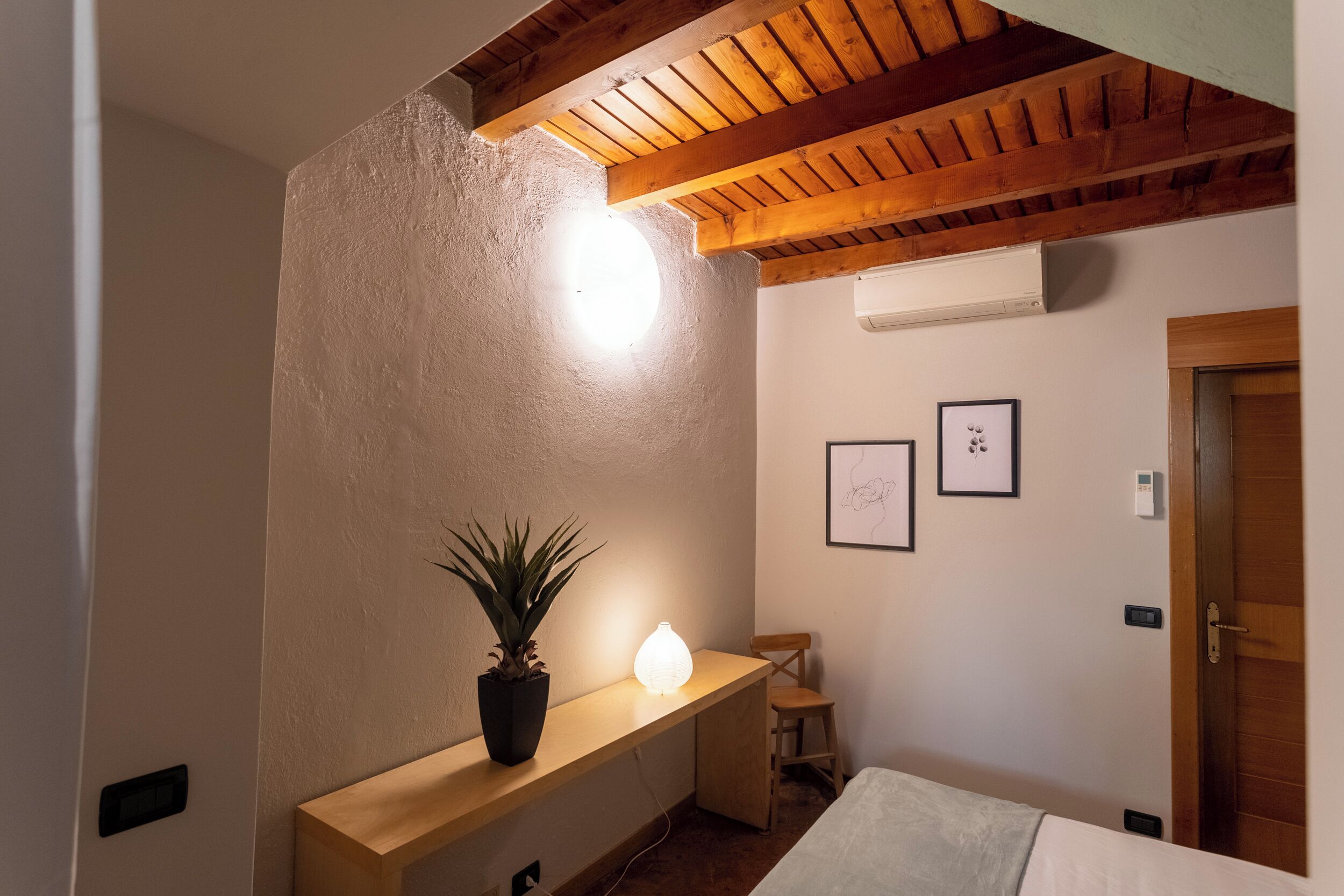 Ein Schlafzimmer mit Holzboden, einem Nachttisch mit Pflanze, einer Wandlampe und einem Bett.