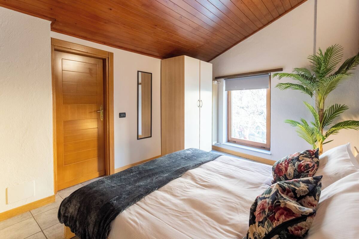 A bedroom with a wooden ceiling, a bed with white bedding, a black throw, and a floral pillow.
