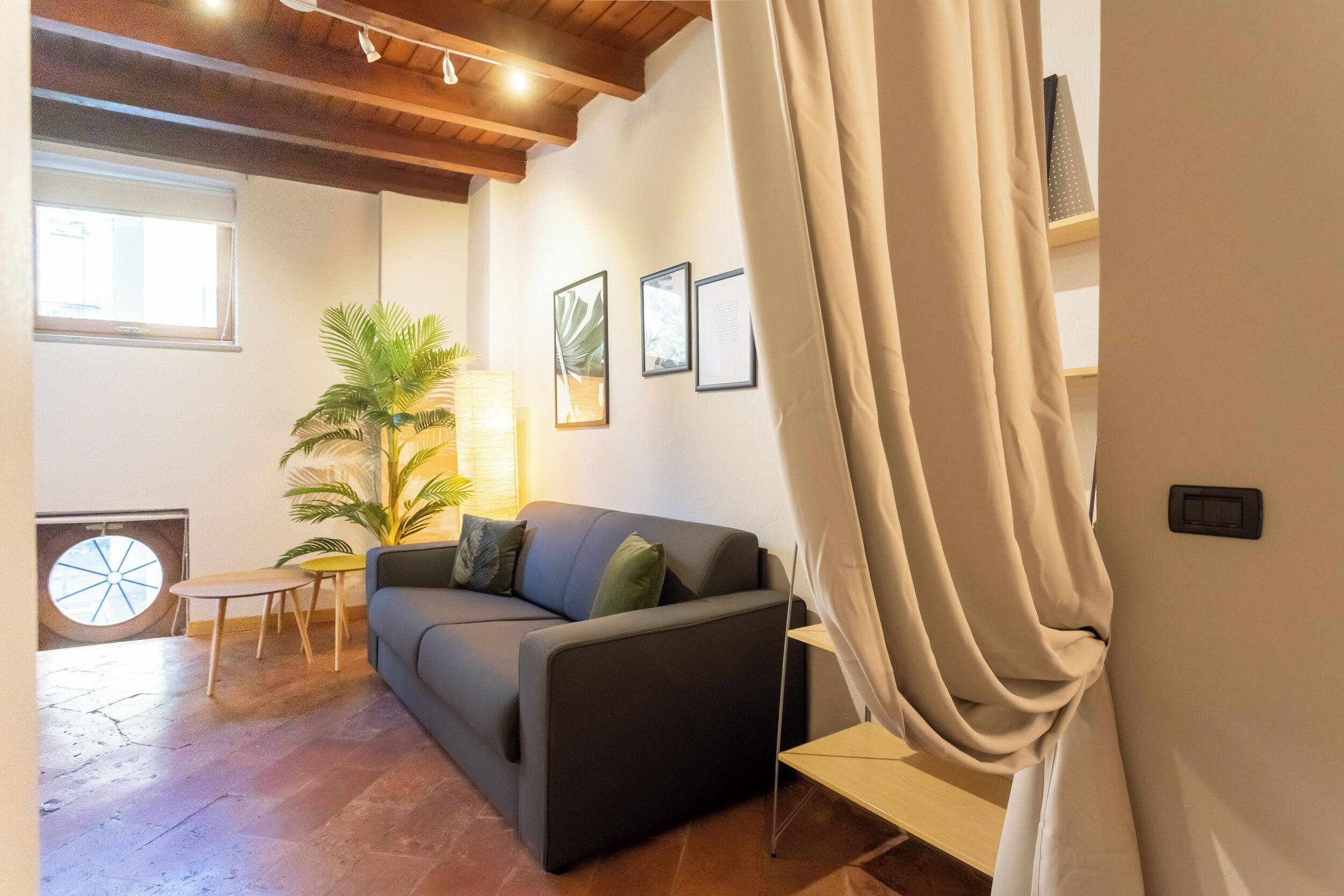 A living room with a sofa, a small table, a potted plant, and a window with blinds.