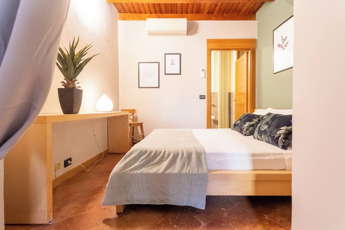 A bedroom with a bed, a desk, a potted plant, and framed pictures on the wall.