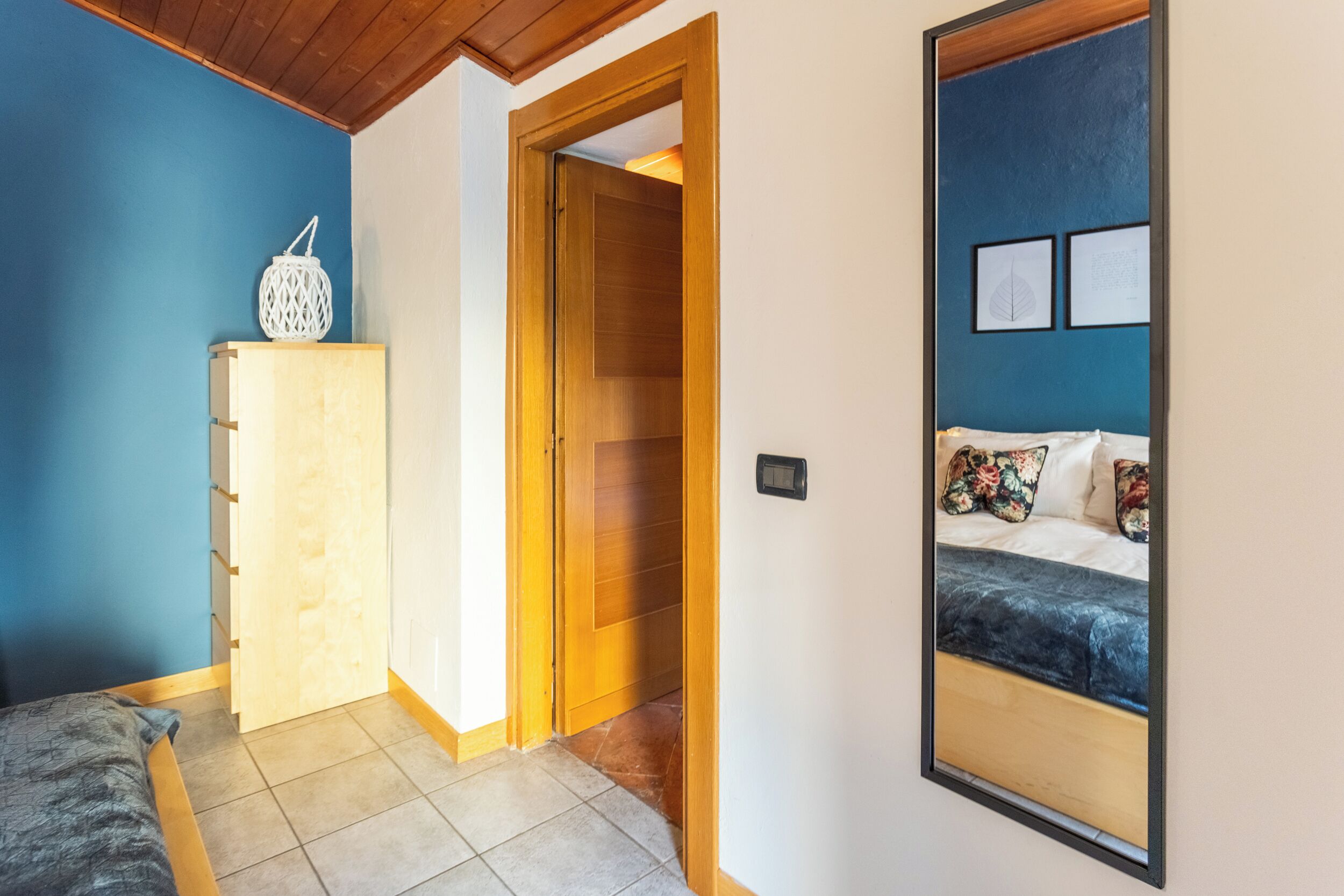 A bedroom with a bed, a wooden wardrobe, a white cabinet, and a mirror reflecting the bed.