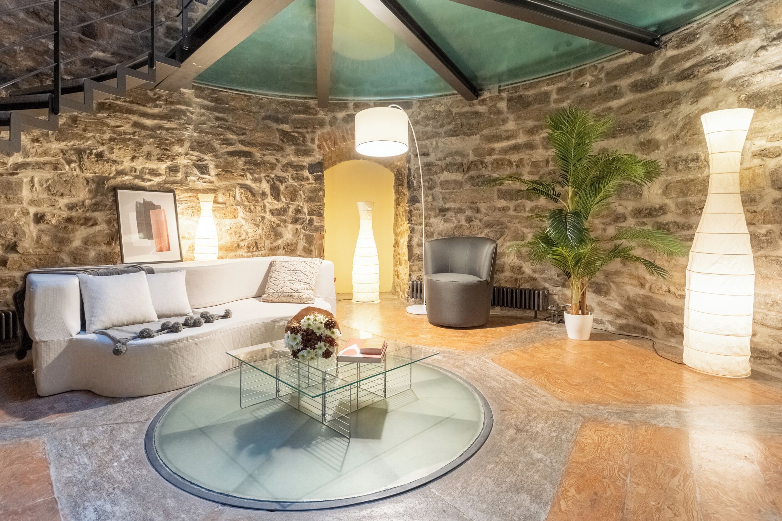 A cozy living area with a stone wall, a round glass coffee table, a white sofa, a gray armchair, and a potted plant.