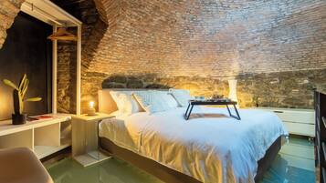 Un dormitorio con techo de ladrillo, cama con ropa de cama blanca, mesita de noche con lámpara y planta colgante.