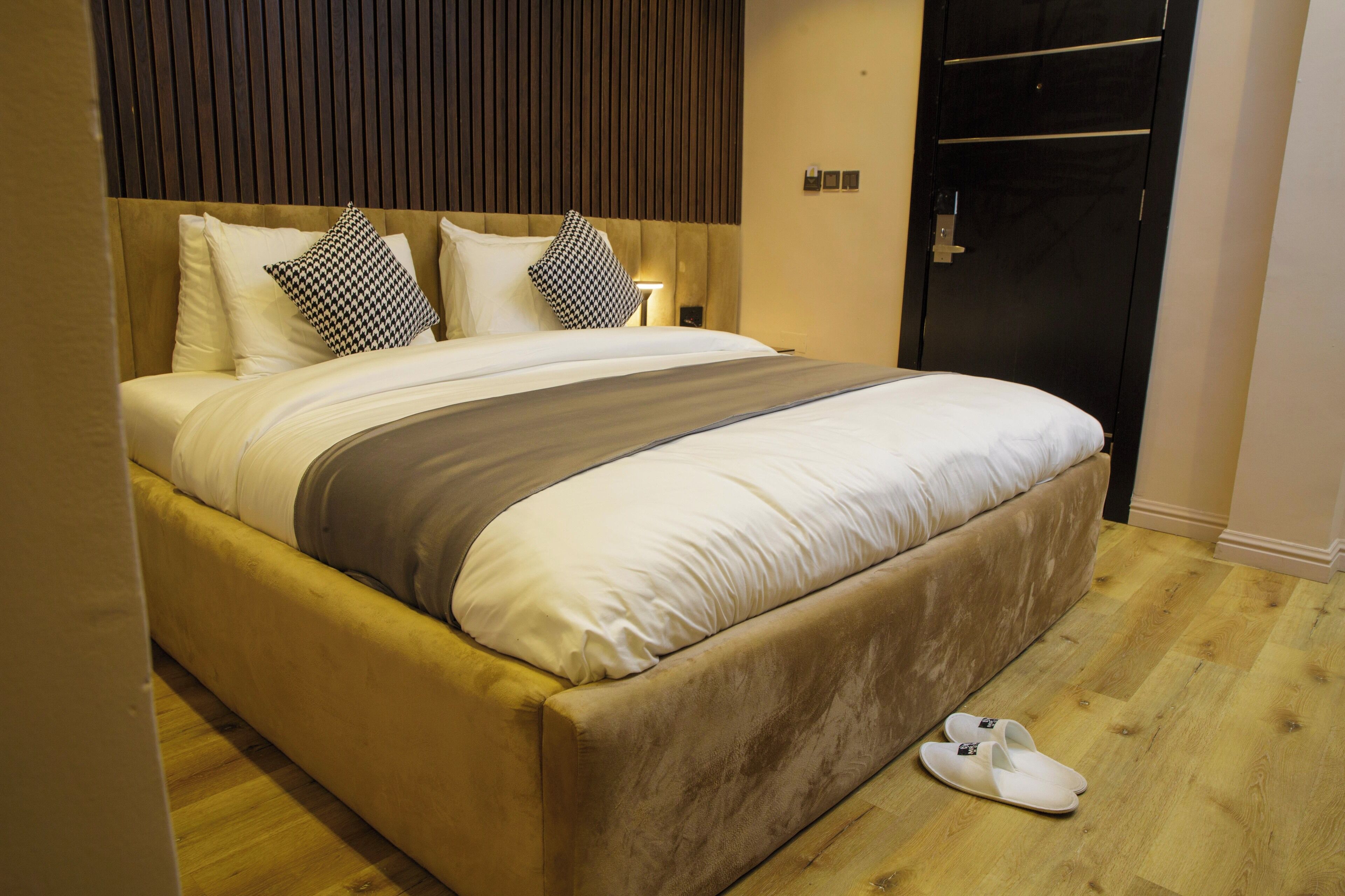 Una cama bien hecha con cabecera, almohadas y manta. Un par de zapatillas están sobre el suelo junto a la cama.