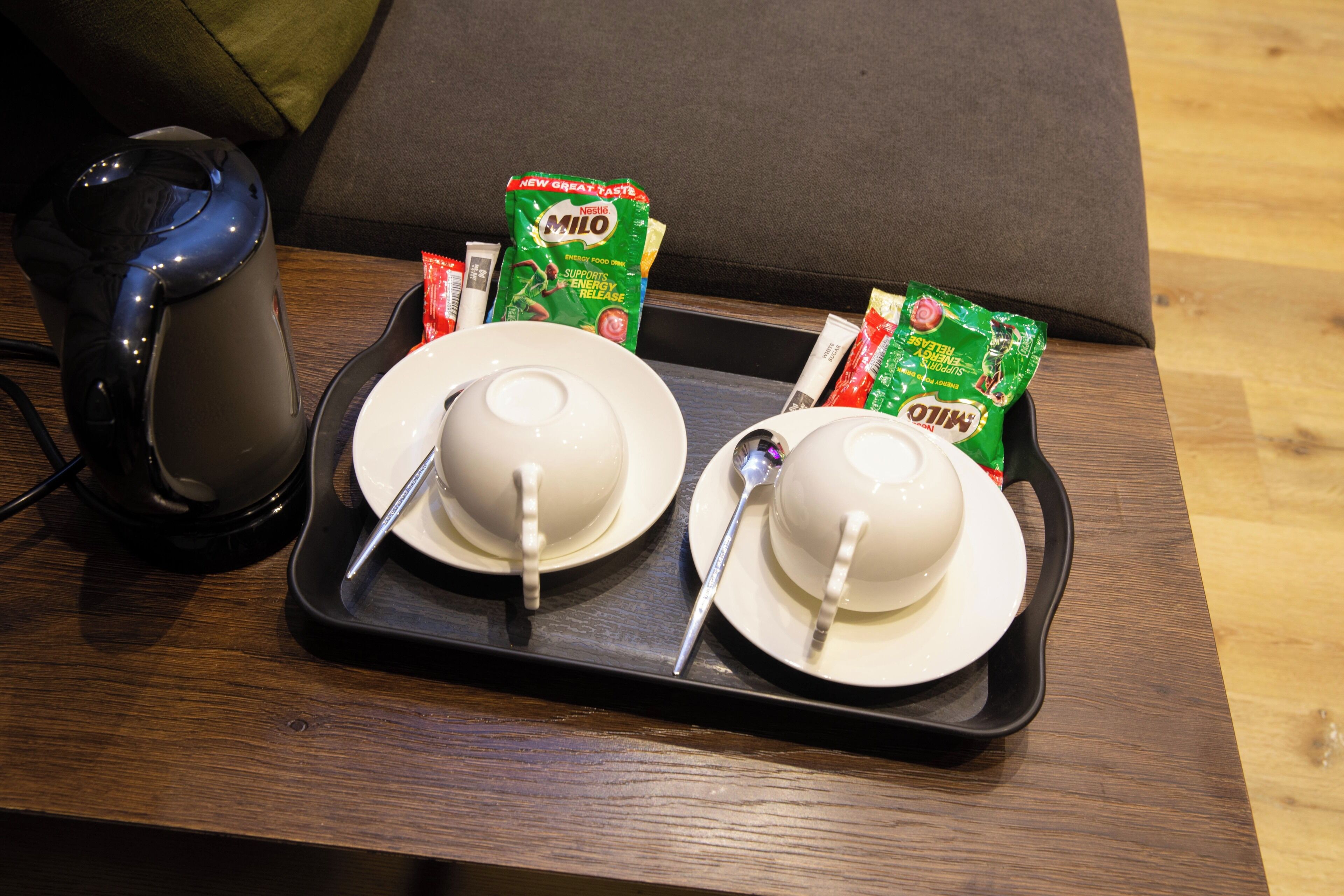 Een dienblad met twee witte bekers, een lepel, een zwarte elektrische waterkoker en pakjes Milo-cereal op een houten tafel.