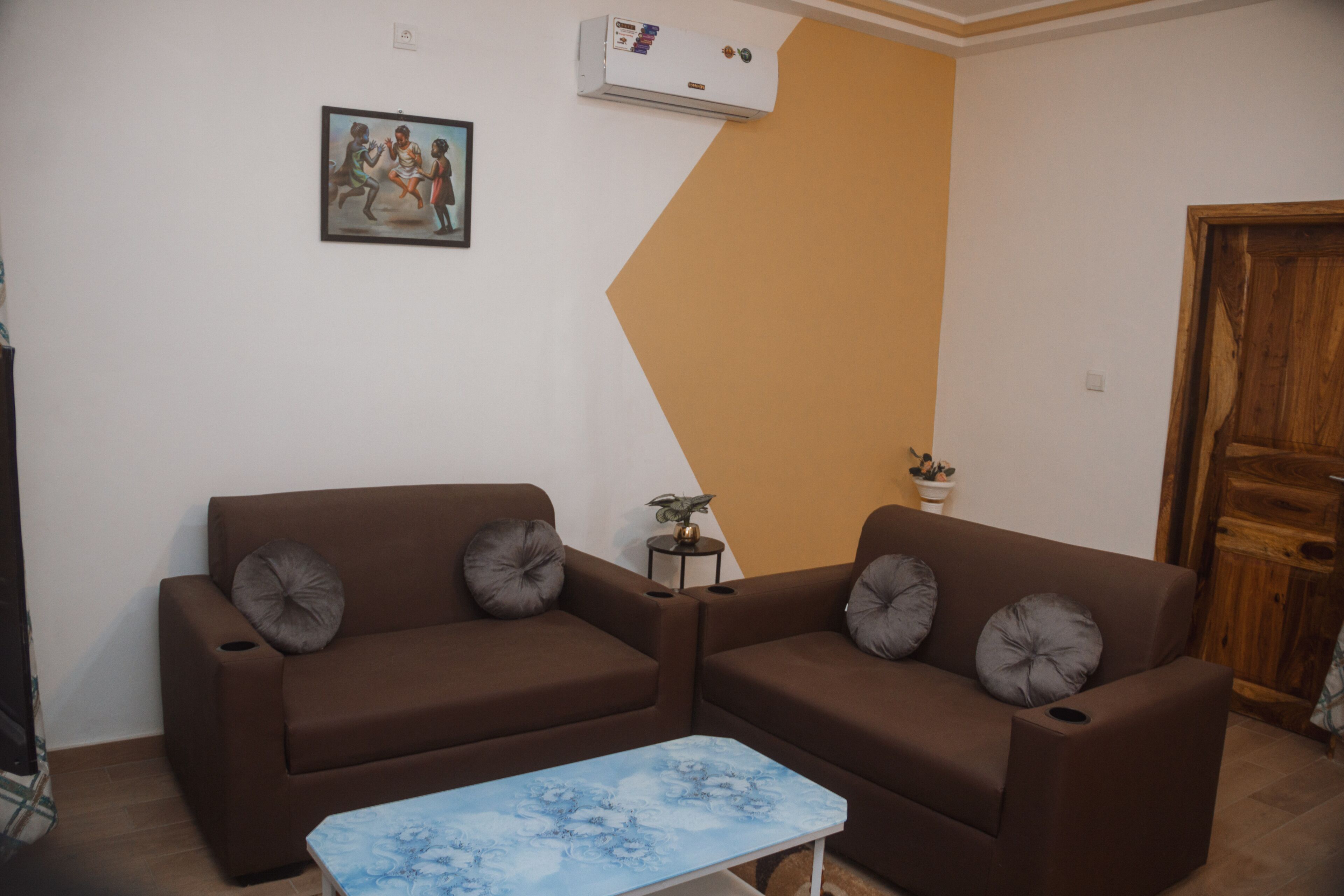 A living room with two brown sofas, a blue coffee table, a wall-mounted air conditioner, a framed picture, and a small plant on a side table.
