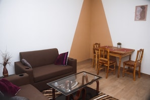 A living room with a brown sofa, a glass coffee table, a dining table with wooden chairs, and a small plant on the wall.