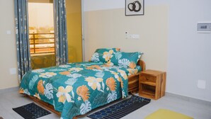 Een slaapkamer met een bed, een houten nachtkastje, een kleine tafel, een raam met gordijnen en een aan de muur hangend schilderij.