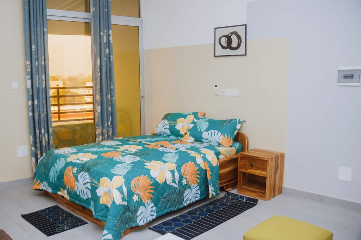 Un dormitorio con una cama, una mesita de noche de madera, una pequeña mesa, una ventana con cortinas y un cuadro colgado en la pared.