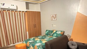 Een kleine kamer met een bed, een slaapbank, een airconditioningapparaat, een gordijn en een lichtschakelaar aan de muur.