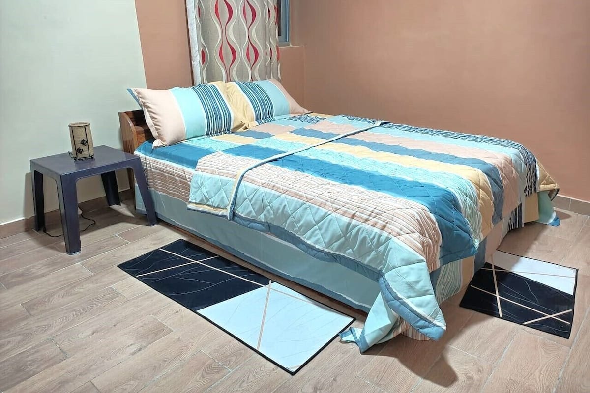 Un dormitorio con cama, mesita de noche, una pequeña mesa y una ventana con cortinas.