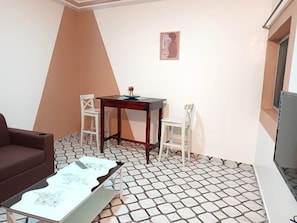 A room with a brown sofa, a dining table with two chairs, a small table with a vase, and a TV on the right.