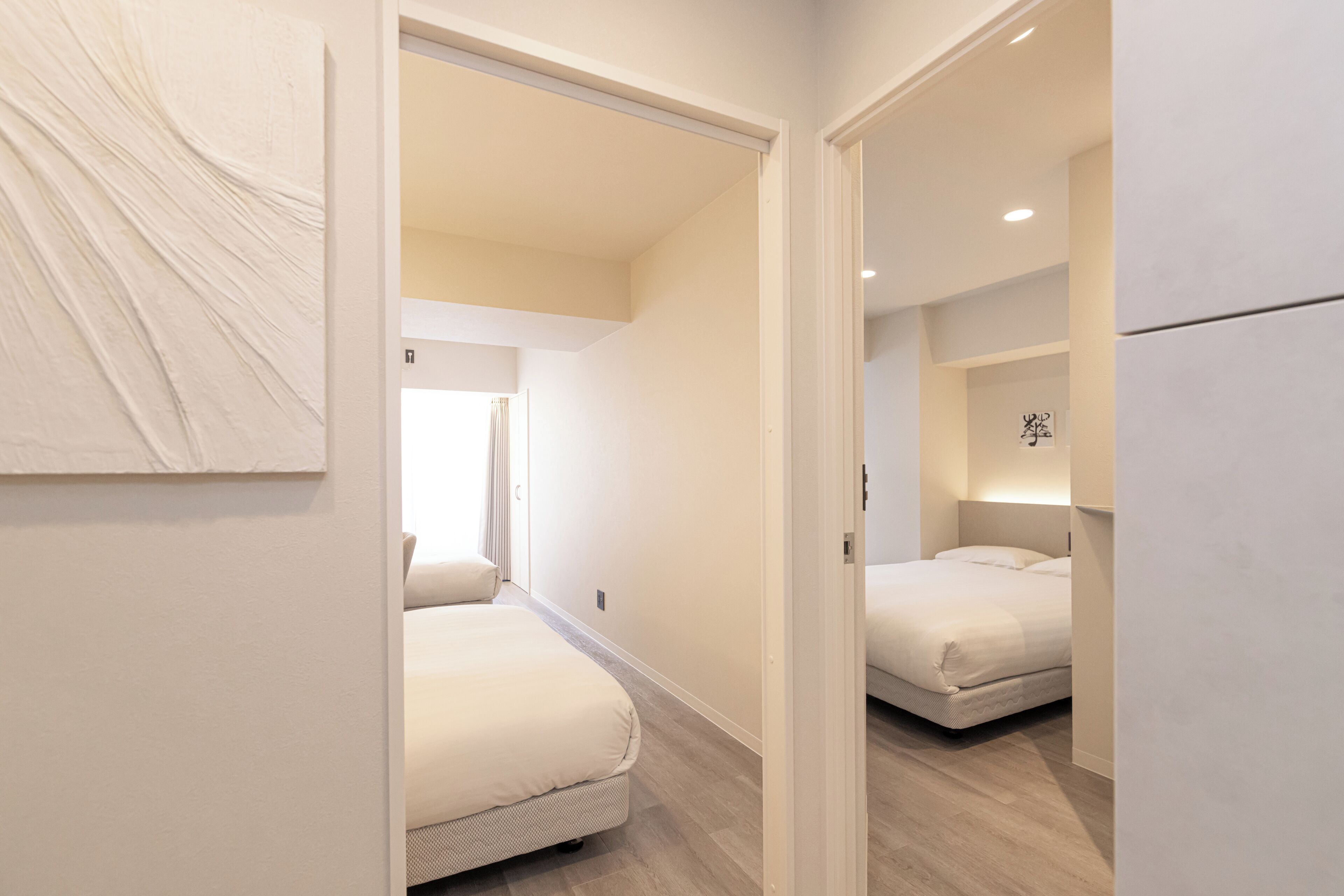 A modern hotel room with a large bed, a small seating area, and a view of the adjacent room.