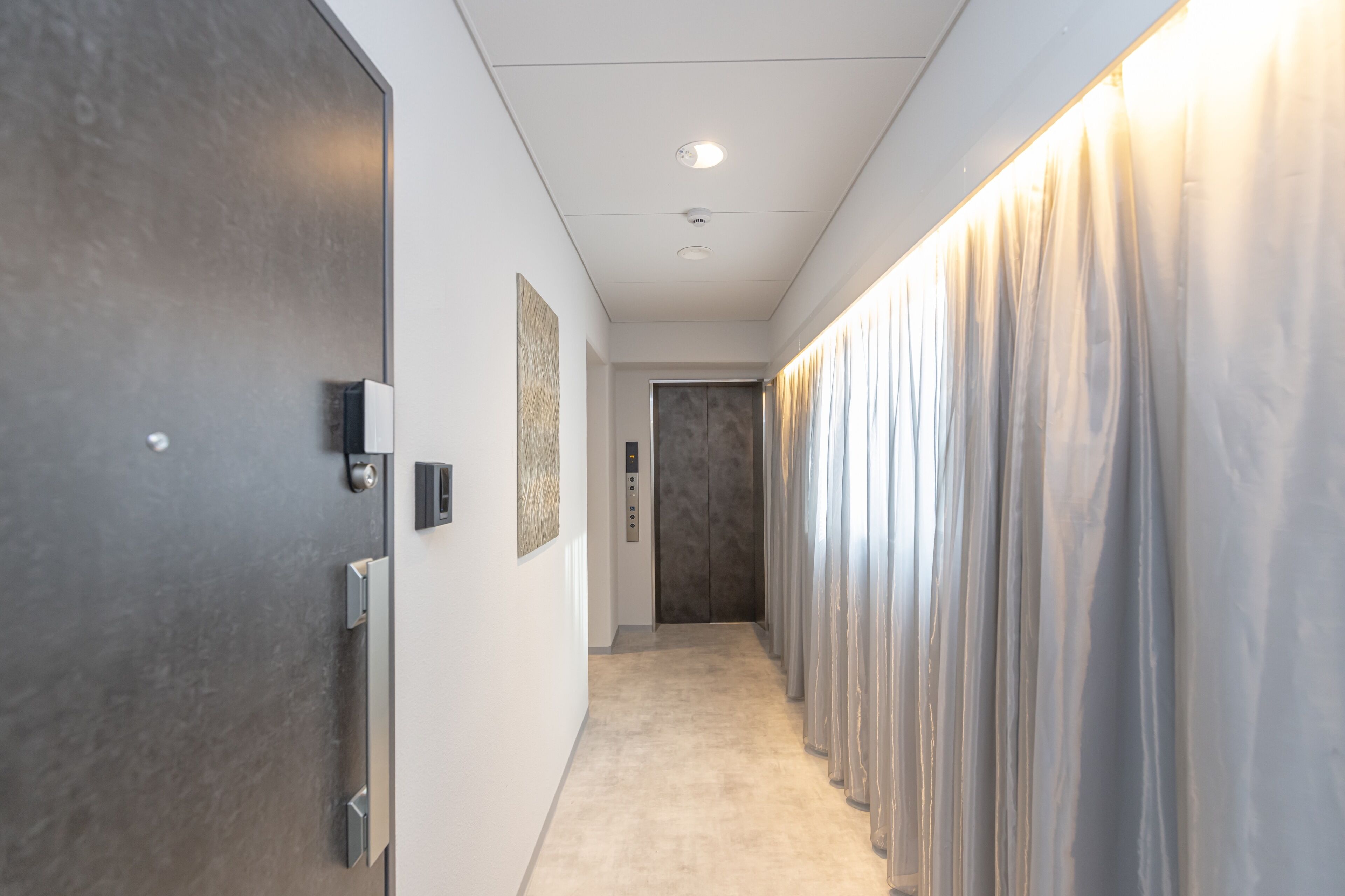 A hallway with a modern elevator, a door with a handle, and a wall-mounted light fixture.