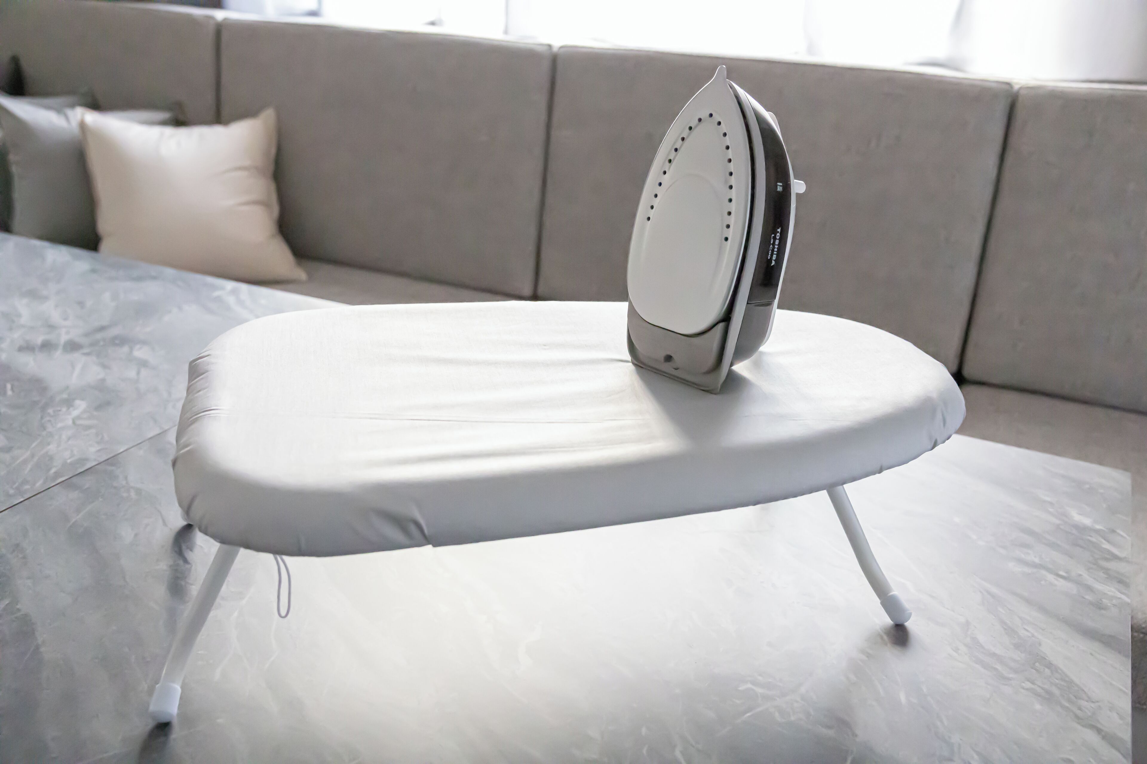 An ironing board with an iron on it, placed on a marble floor in a room with a grey sofa and a white pillow in the background.