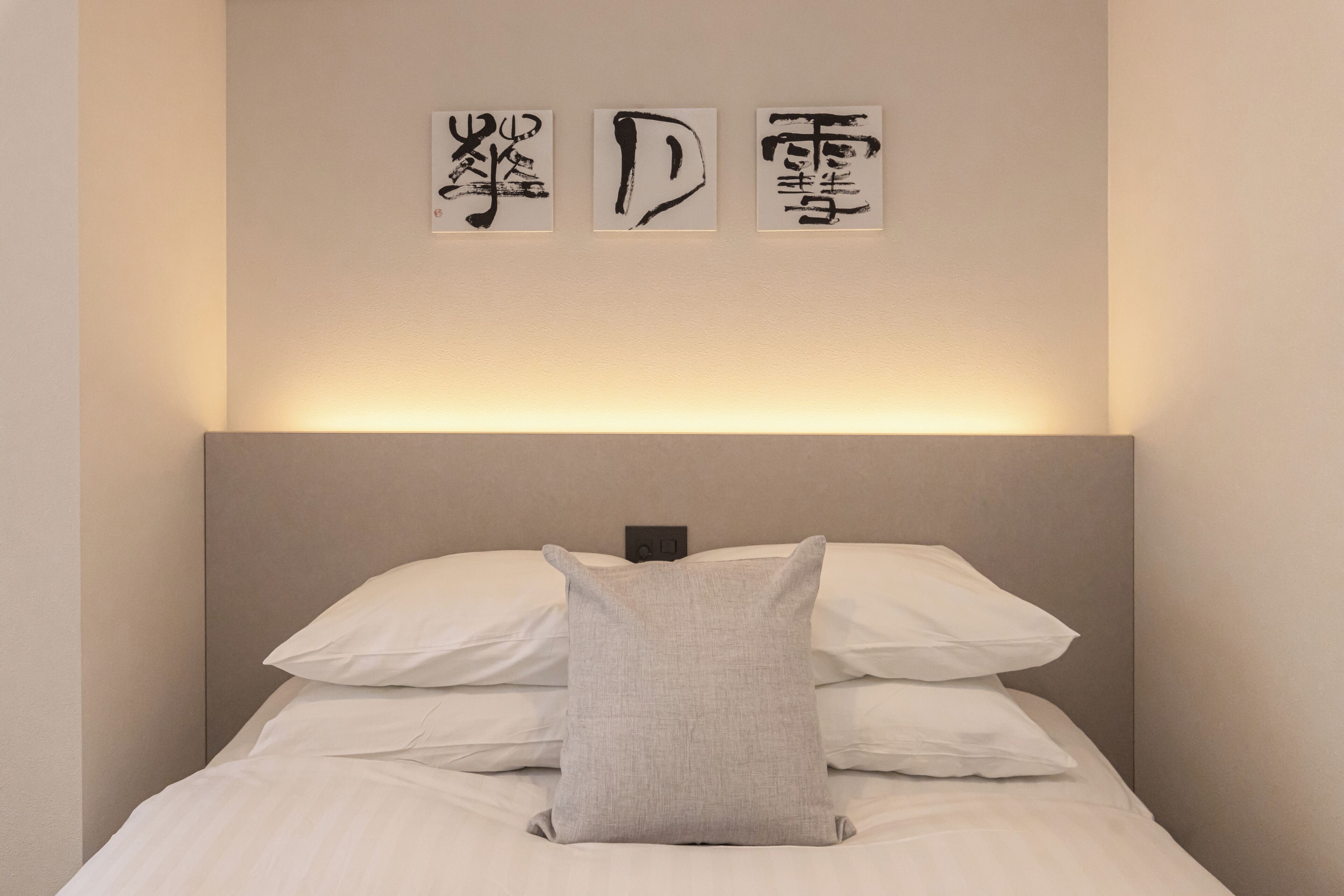 A neatly made bed with white linens and a single grey pillow, a wall-mounted light above, and three black calligraphy pieces on the wall.