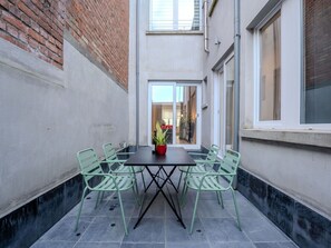 A narrow outdoor patio with a table and chairs, a potted plant, and a view into the adjacent building.