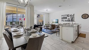 Eine moderne Küche mit einer zentralen Kochinsel, ein Essbereich mit Tisch für vier Personen und ein Wohnbereich mit Sofa und Couchtisch.