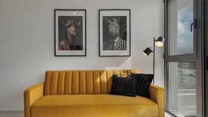 En soffa i senapsgul färg med svarta kuddar, två inramade porträtt av djur på väggen och en golvlampa.