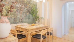 A dining area with a wooden table set for four, surrounded by chairs. A large vase with flowers is on the table, and a mural of a tree and birds is on the wall behind.
