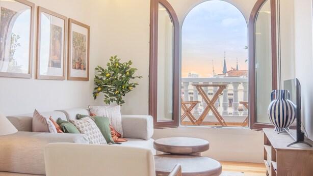 A living room with a sofa, a small round table, a potted plant, and a view of a cityscape through arched windows.