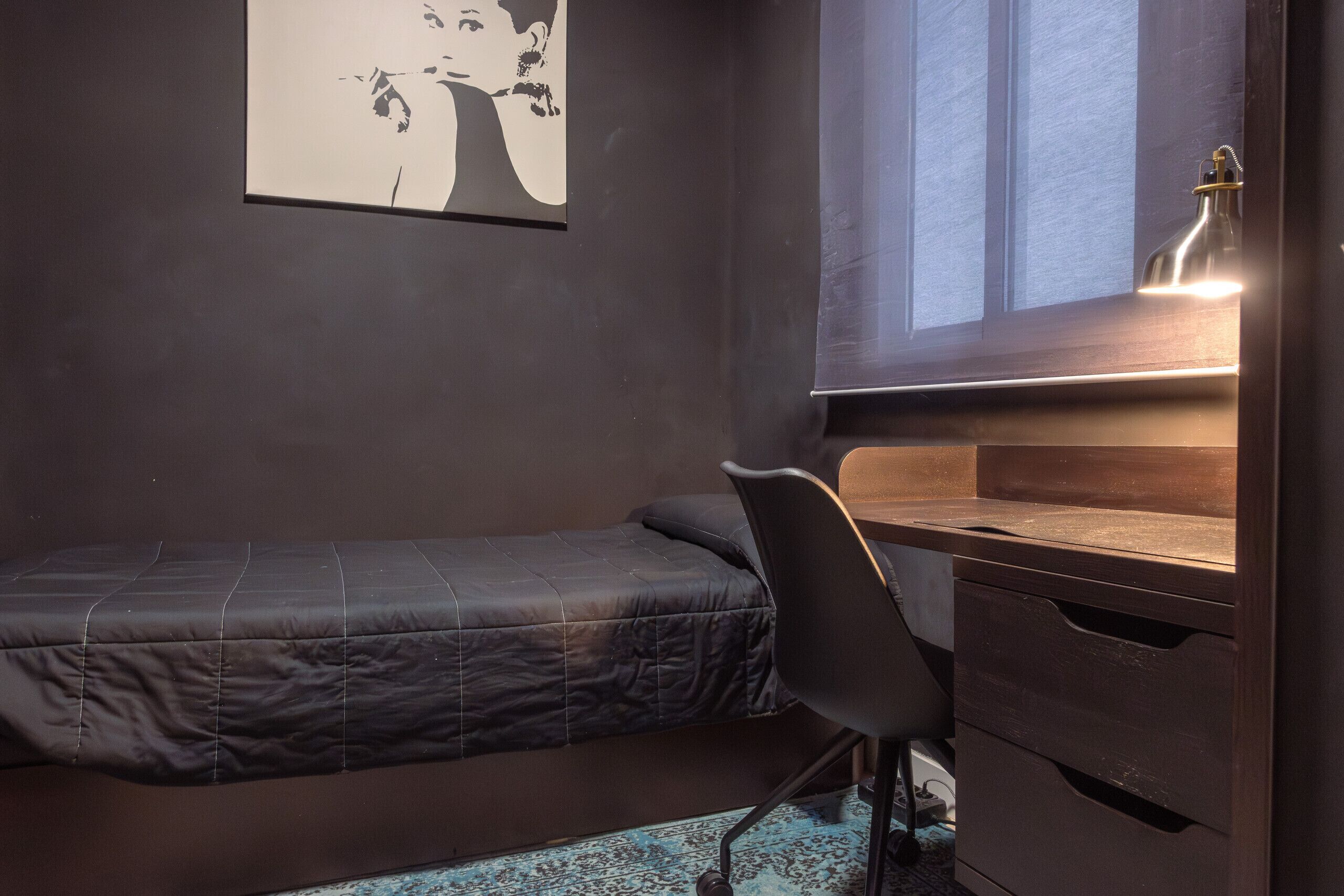 A small bedroom with a single bed, a desk with a lamp, and a chair.