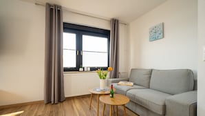 En moderne stue med en grå sofa, et lille træbord med en flaske og en vase med blomster, samt et vindue med gardiner.