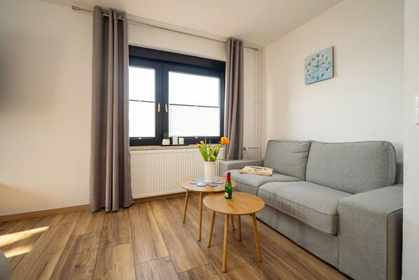 Una sala de estar moderna con un sofá gris, una mesita de madera con una botella y un jarrón de flores, y una ventana con cortinas.