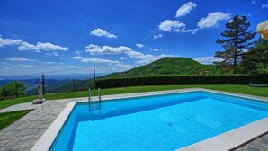 Une piscine rectangulaire bordée de pierres, entourée d’un sentier également en pierre, le tout se fondant dans un paysage verdoyant avec une colline luxuriante en arrière-plan et un ciel bleu dégagé.