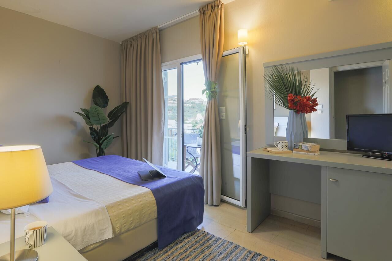 A hotel room with a bed, a desk with a computer, a TV, a window with curtains, and a plant.