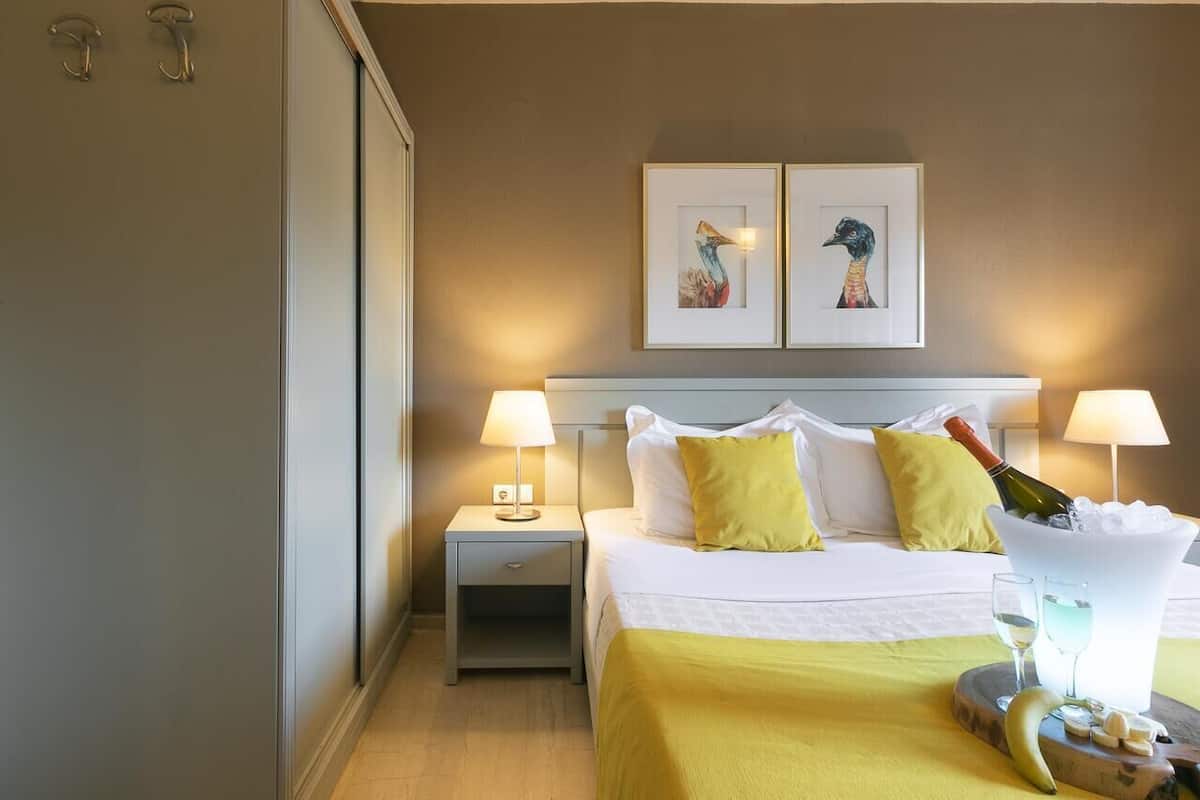 A hotel room with a bed, two bedside lamps, a nightstand, a framed artwork above the bed, and a bucket with a bottle and glasses on the bed.