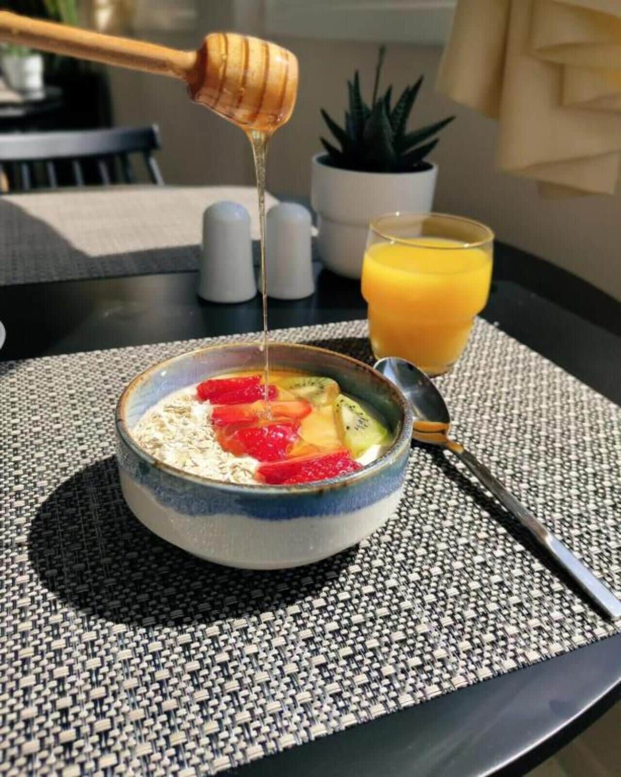A bowl of cereal with fruits and honey being poured, a glass of orange juice, and a spoon on a table with a woven placemat.